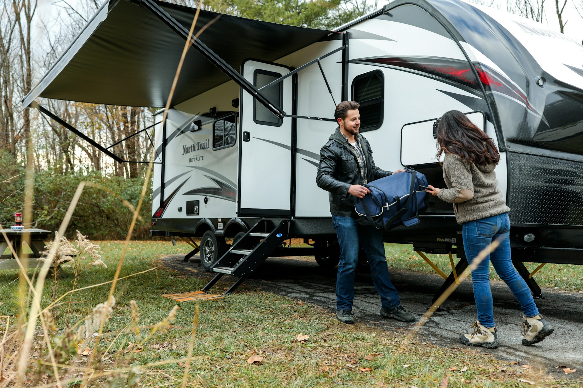 Two campers unloading a bag beside a travel trailer with its awning extended